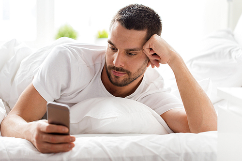 technology, internet, communication and people concept - happy young man texting on smartphone in bed at home in morning