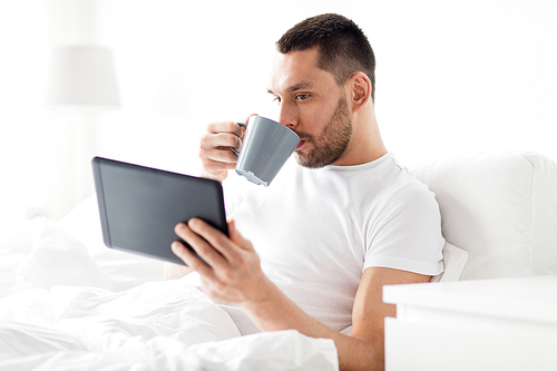 technology, internet, communication and people concept - young man with tablet pc computer drinking coffee in bed at home bedroom