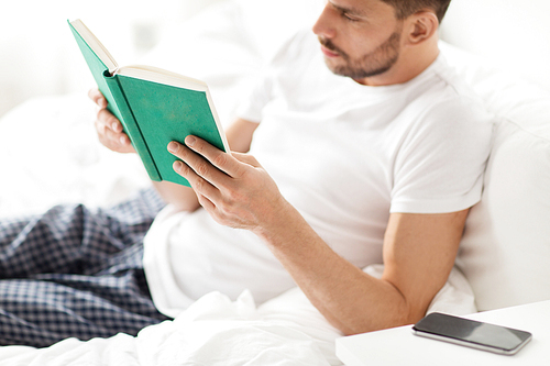 people and rest concept - man reading book in bed at home