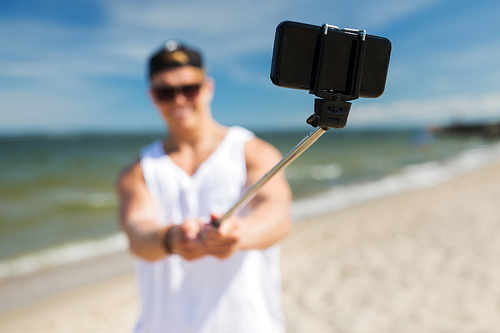 technology, summer holidays and people concept - happy young man taking picture by smartphone on selfie stick on beach