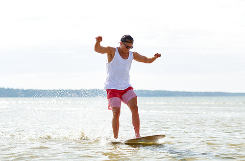 skimboarding, water sport and people concept - happy young man riding on skimboard on summer beach
