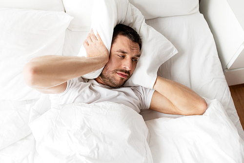people, bedtime and rest concept - man lying in bed with pillow suffering from noise at home
