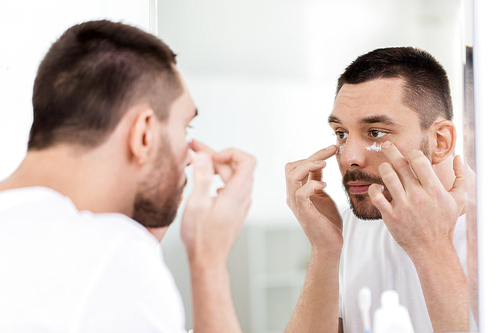 grooming, skin care and people concept - young man applying cream to face and looking to mirror at home bathroom