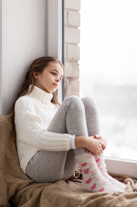 childhood, sadness and people concept - sad beautiful girl in sweater sitting on sill at home window in winter