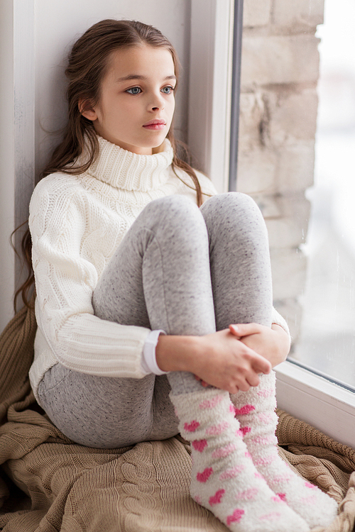 childhood, sadness and people concept - sad beautiful girl in sweater sitting on sill at home window in winter
