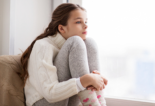 childhood, sadness and people concept - sad beautiful girl in sweater sitting on sill at home window in winter