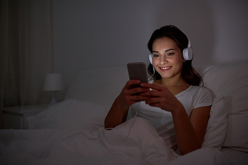 technology, leisure and people concept - happy smiling young woman with smartphone and headphones listening to music in bed at night