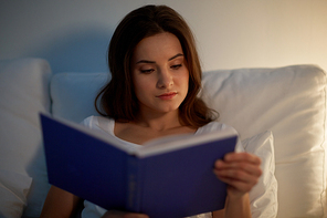 leisure and people concept - young woman reading book in bed at night home