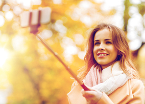 season, technology and people concept - beautiful young happy woman taking picture with smartphone selfie stick in autumn park