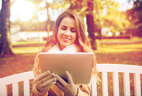 season, technology and people concept - beautiful happy young woman with tablet pc computer sitting on bench in autumn park