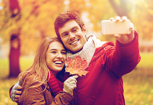 love, technology, relationship, family and people concept - smiling couple with maple leaf taking selfie by smartphone in autumn park