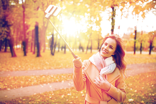 season, technology and people concept - beautiful young happy woman taking picture with smartphone selfie stick in autumn park