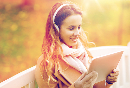 season, technology and people concept - beautiful happy young woman with tablet pc computer and headphones listening to music and sitting on bench in autumn park