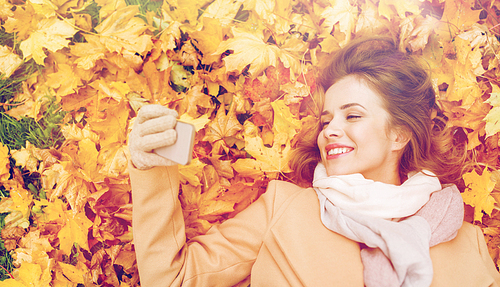 season, technology and people concept - beautiful young woman lying on ground and autumn leaves and taking selfie with smartphone
