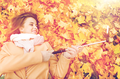 season, technology and people concept - beautiful young woman lying on ground and autumn leaves and taking picture with smartphone selfie stick