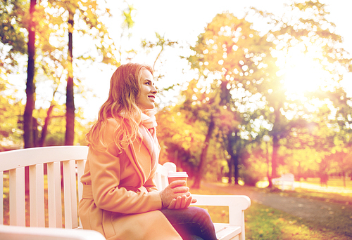 season, hot drinks and people concept - beautiful happy young woman drinking coffee or tea from disposable paper cup sitting on bench in autumn park