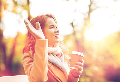 season, hot drinks and people concept - beautiful happy young woman drinking coffee or tea from disposable paper cup and waving hand in autumn park