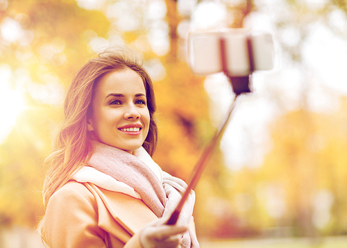 season, technology and people concept - beautiful young happy woman taking picture with smartphone selfie stick in autumn park