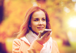 season, technology and people concept - beautiful young woman walking in autumn park and using voice command recorder on smartphone