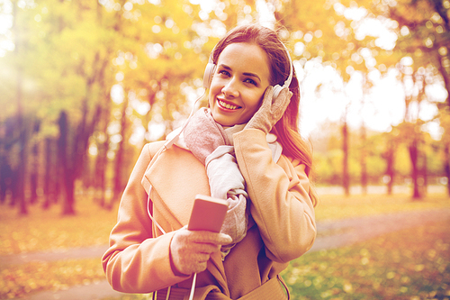 season, technology and people concept - beautiful happy young woman with headphones listening to music on smartphone walking in autumn park