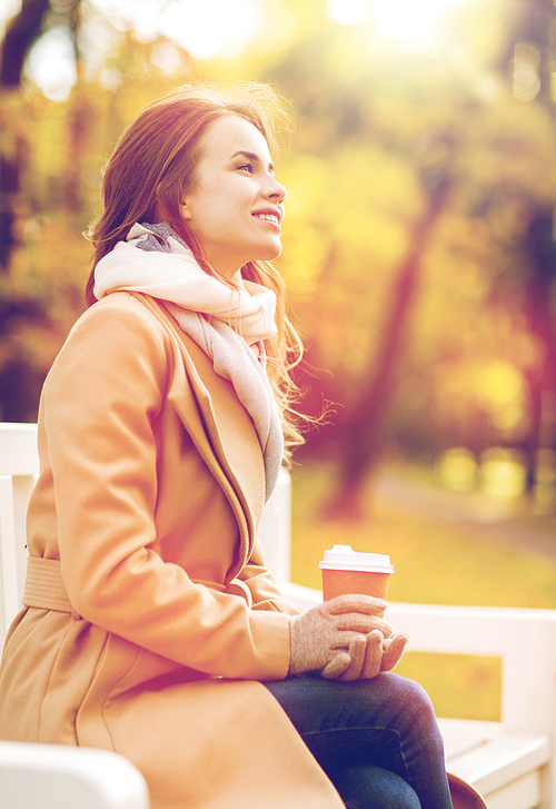season, hot drinks and people concept - beautiful happy young woman drinking coffee or tea from disposable paper cup sitting on bench in autumn park