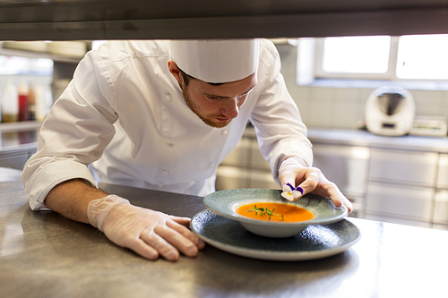 food cooking, profession and people concept - happy male chef cook serving and decorating plate of soup with pansy flower at restaurant kitchen