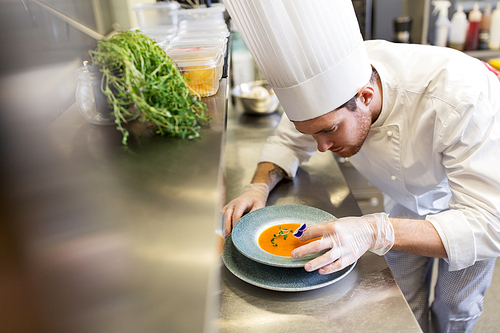 food cooking, profession and people concept - happy male chef cook serving and decorating plate of soup with pansy flower at restaurant kitchen