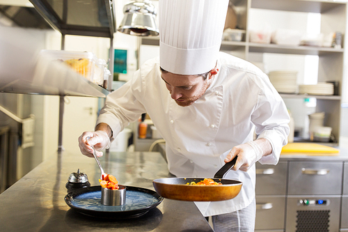food cooking, profession and people concept - happy male chef cook with frying pan and spoon serving stewed vegetables on plate at restaurant kitchen