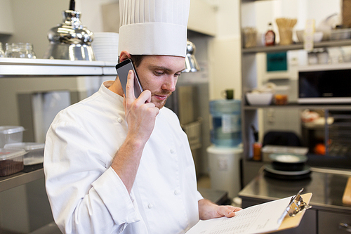 cooking, profession and people concept - male chef cook with clipboard calling on smartphone at restaurant kitchen