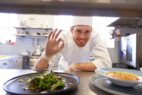 food cooking, profession and people concept - happy male chef cook with plate of soup and salad at restaurant kitchen table showing ok hand sign