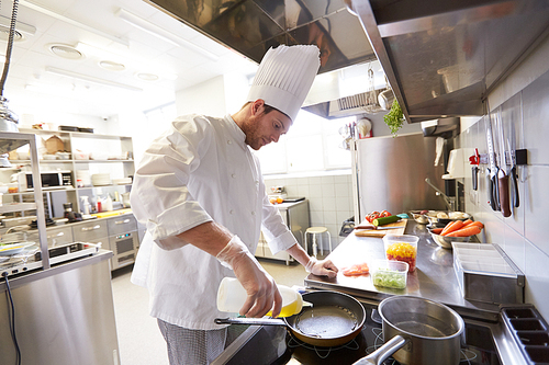 cooking food, profession and people concept - happy male chef cook pouring oil to frying pan at restaurant kitchen
