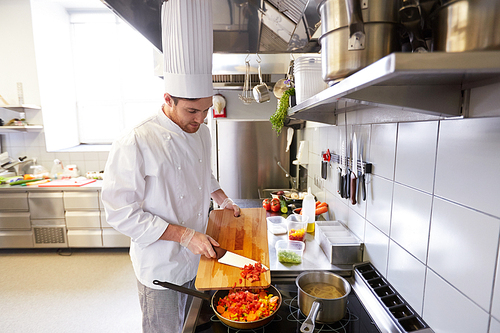 cooking food, profession and people concept - male chef cook with knife and cutting board adding chopped tomatoes to frying pan at restaurant kitchen