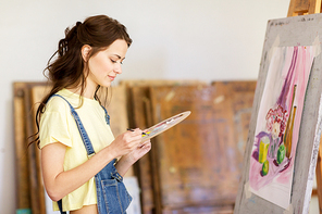 art school, creativity and people concept - student girl or young woman artist with easel, palette and paint brush painting still life picture at studio