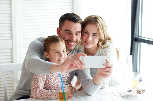 family, parenthood, technology and people concept - happy mother, father and little girl having dinner and taking selfie by smartphone at restaurant