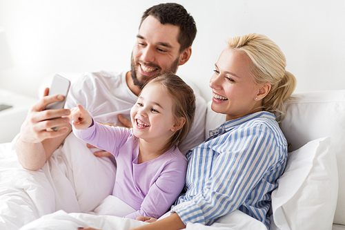 people, family and technology concept - happy mother, father and little girl taking selfie with smartphone in bed at home