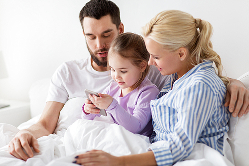 people, family and technology concept - happy mother, father and little girl with smartphone in bed at home