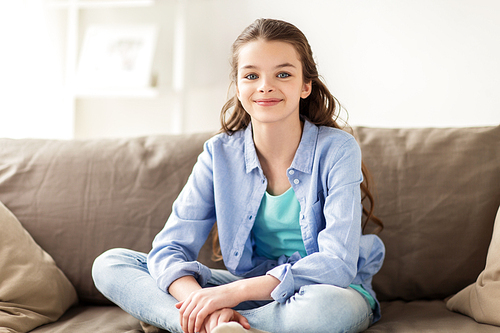 people and childhood concept - happy smiling preteen girl sitting on sofa at home