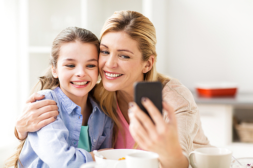 family, technology and people concept - happy mother and daughter with smartphone having breakfast and taking selfie at home kitchen