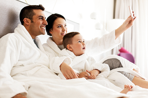 people, family and technology concept - happy mother, father and little boy in bathrobe taking selfie by smartphone in bed or hotel room