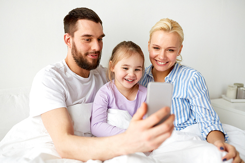 people, family and technology concept - happy mother, father and little girl taking selfie with smartphone in bed at home