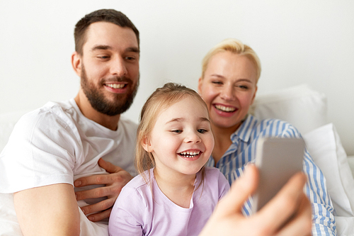 people, family and technology concept - happy mother, father and little girl taking selfie with smartphone in bed at home