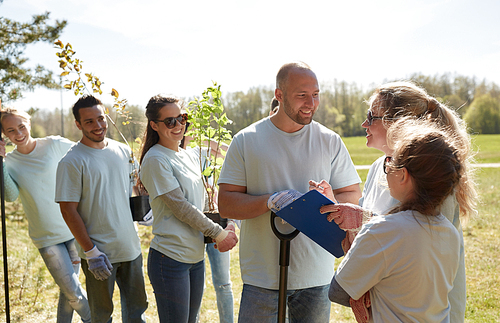 volunteering, charity, people and ecology concept - group of happy volunteers with tree seedlings and clipboard talking in park