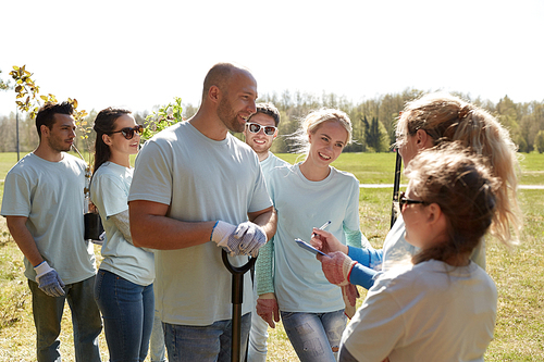 volunteering, charity, people and ecology concept - group of happy volunteers with tree seedlings and clipboard talking in park