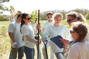 volunteering, charity, people and ecology concept - group of happy volunteers with tree seedlings and clipboard in park