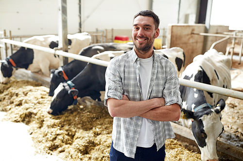 agriculture industry, farming, people and animal husbandry concept - happy smiling young man or farmer with herd of cows in cowshed on dairy farm