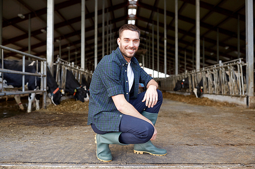 agriculture industry, farming, people and animal husbandry concept - happy smiling young man or farmer with herd of cows in cowshed on dairy farm