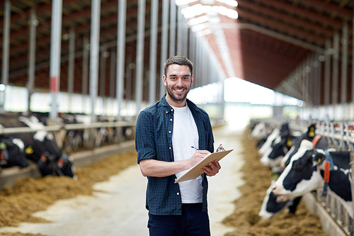 agriculture industry, farming, people and animal husbandry concept - happy smiling young man or farmer with clipboard and cows in cowshed on dairy farm