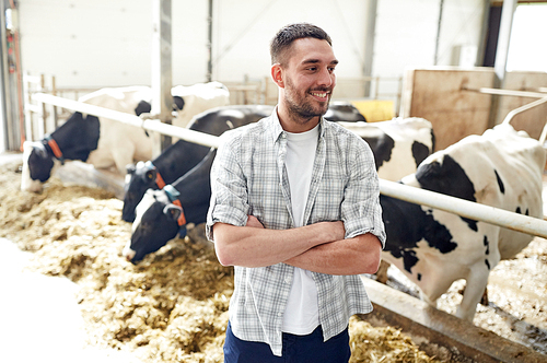 agriculture industry, farming, people and animal husbandry concept - happy smiling young man or farmer with herd of cows in cowshed on dairy farm