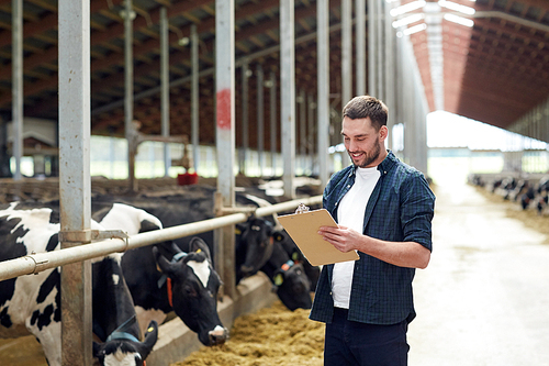 agriculture industry, farming, people and animal husbandry concept - happy smiling young man or farmer with clipboard and cows in cowshed on dairy farm