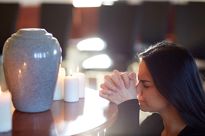 people and mourning concept - sad woman with cinerary urn and candles praying at funeral in church